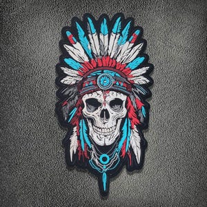 Può includere: Una toppa ricamata raffigurante un teschio umano che indossa un copricapo di piume. Il design utilizza fili di colore bianco, rosso e turchese. Il teschio è centrato, ornato da una collana di perline e incorniciato da un bordo nero su sfondo ruvido.
