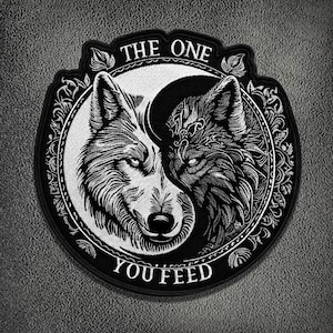 Op de afbeelding: Een ronde geborduurde patch met een zwart-wit yin-yang ontwerp bestaande uit twee wolvenkoppen die naar elkaar toe kijken. De tekst THE ONE staat in een boog boven de wolven en YOU FEED eronder, alles binnen een decoratieve rand.