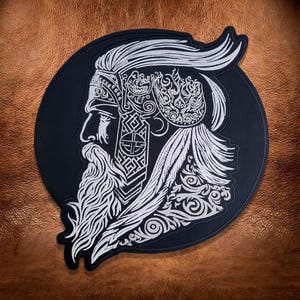 May include: A circular black embroidered patch featuring a white thread design of a profile view of a bearded figure with long hair, wearing an ornate headpiece and decorative armor with intricate geometric and swirling patterns throughout.