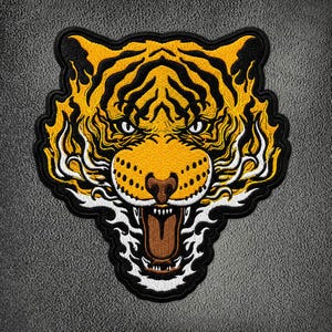 Peut inclure: Un écusson brodé représentant une tête de tigre avec un pelage jaune, des rayures noires et des accents blancs. Le tigre est représenté la gueule ouverte montrant ses dents et une langue brune, entouré de motifs de flammes blancs et jaunes.