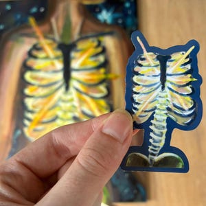 Op de afbeelding: Een gestanste sticker met een illustratie van een menselijke ribbenkast en ruggengraat tegen een donkerblauwe achtergrond. De botten zijn weergegeven in tinten geel, wit en oranje, met een zwart borstbeen. De sticker weerspiegelt een groter schilderij.