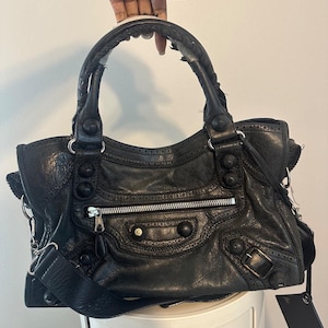 May include: A black leather handbag featuring a textured finish, dual top handles with braided detailing, a front zippered pocket, and decorative metal studs. The bag includes a detachable shoulder strap and a hanging leather luggage tag.