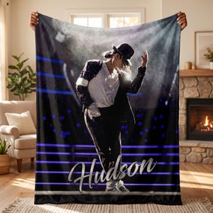 May include: A soft fleece blanket featuring a photographic print of a performer wearing a black sequined jacket, white shirt, black trousers, and a fedora hat. The background shows a stage with blue horizontal light beams. The name Hudson is printed.