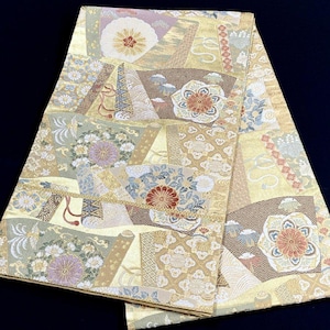 Könnte beinhalten: Ein rechteckiges Textil mit goldfarbener Basis und einem komplexen Muster aus überlappenden Fächerformen. Das Design enthält detaillierte Blumenmotive, darunter Chrysanthemen, in Weiß-, Violett-, Rot- und Blautönen mit metallischen Akzenten.