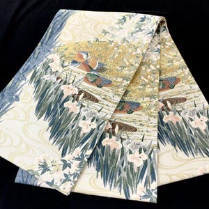 May include: A folded textile featuring a cream-colored background with a detailed woven pattern of mandarin ducks swimming in water. The design includes gold metallic accents, green reeds, and light pink iris flowers arranged in a repeating motif.