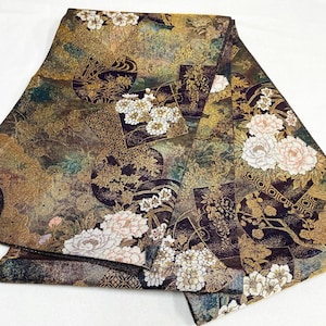 May include: A folded textile featuring a dark background with intricate gold metallic patterns, including geometric shapes and floral motifs. The fabric displays clusters of white and pale pink flowers, with detailed gold leaf-like textures.