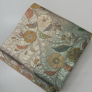 May include: A folded textile featuring an intricate woven floral pattern in shades of muted gold, sage green, and soft brown. The fabric displays a dense arrangement of blossoms and leaves with a subtle sheen, creating a textured surface appearance.