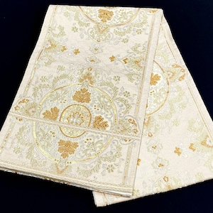 May include: A long, rectangular textile runner featuring a cream-colored base with intricate, woven patterns in metallic gold and silver thread. The design includes symmetrical floral motifs, scrolling vines, and circular medallions.