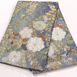 May include: A folded textile featuring a blue-grey base with an intricate woven pattern of white floral blossoms and golden fern-like foliage. The fabric has a textured, shimmering finish with subtle accents of green and copper throughout the design.