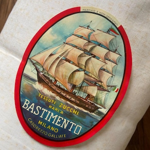 Può includere: Un rotolo di tessuto color avorio con una grande etichetta ovale dal bordo rosso. L'etichetta raffigura un veliero a più alberi su acqua blu e riporta il testo: MARCHIO E NOMI DEPOSITATI, TESSUTI ZUCCHI MARCA BASTIMENTO MILANO CASOREZZO GALLIATE.