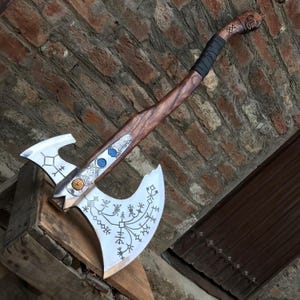 Puede incluir: Un hacha de doble filo con una cabeza de metal plateado pulido que presenta intrincados símbolos rúnicos grabados en negro. El mango está hecho de madera marrón oscura con una empuñadura envuelta en negro y un pomo tallado, acentuado por incrustaciones azules y doradas.