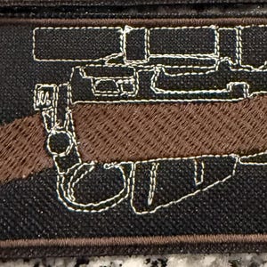 Puede incluir: Un parche rectangular bordado que presenta una culata de rifle marrón sobre un fondo negro. El diseño incluye un contorno blanco cosido de una mira telescópica y un mecanismo de gatillo, con un borde marrón oscuro que enmarca la pieza.