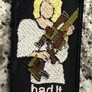 May include: A rectangular embroidered patch with a black background featuring a blonde figure in a white top holding a brown rifle. The text Jolene is embroidered in white at the top, and the text had it comin... is embroidered in white at the bottom.