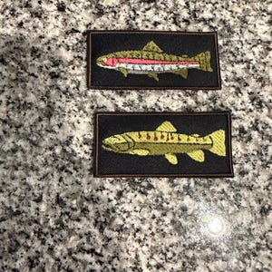 May include: Two rectangular embroidered patches with black borders. The top patch features a rainbow trout with a pink lateral stripe on a black background. The bottom patch displays a yellow and green fish with red spots on a black background.