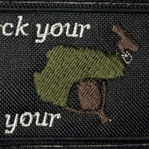 May include: A rectangular black embroidered patch featuring the white text "Check your gear" at the top and "and your ego." at the bottom. In the center, an embroidered graphic depicts a brown firearm with a green camouflage-patterned cover.