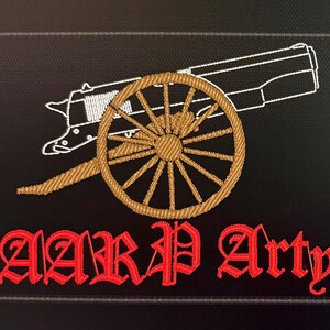 May include: A rectangular black embroidered patch featuring a white outline of a cannon with a gold-colored wheel. Below the cannon, the text "AARP Arty" is stitched in a bold, red, gothic-style font against the black fabric background.