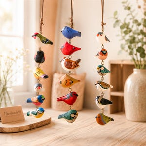 May include: Three vertical hanging ornaments featuring carved wooden birds in various colors including red, blue, yellow, and green. Each ornament consists of a leather cord attached to a metal chain with multiple small, hand-painted bird figures.
