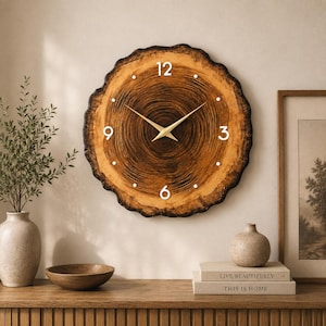 Peut inclure: Une horloge murale ronde fabriquée à partir d'une tranche de bois naturel avec des cernes de croissance brun foncé visibles. Le cadran présente des chiffres blancs à 12, 3, 6 et 9, des points blancs pour les autres heures et des aiguilles dorées.