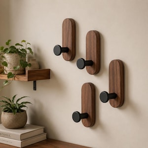 Puede incluir: Cuatro ganchos de pared, cada uno con una base vertical de madera oscura de forma rectangular redondeada y una clavija cilíndrica de color negro mate. Los ganchos están dispuestos en un patrón escalonado sobre una pared clara.