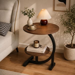 Peut inclure: Une table d'appoint ronde à deux niveaux avec une finition en bois foncé et un cadre en métal noir en forme de C. Le plateau supérieur porte une lampe plissée, un petit vase avec de la verdure et une tasse. L'étagère inférieure porte une bougie et un livre.