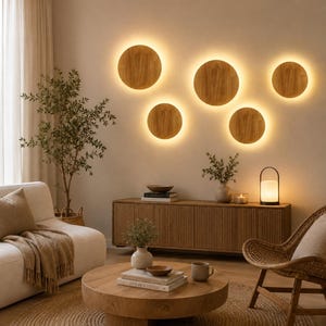 Op de afbeelding: Vijf ronde houten wandlampen met warme achtergrondverlichting gemonteerd aan een muur. De kamer bevat een lichtgekleurde bank, een houten salontafel met boeken, een houten dressoir met latten, een rotan stoel en een draagbare lamp met kap.
