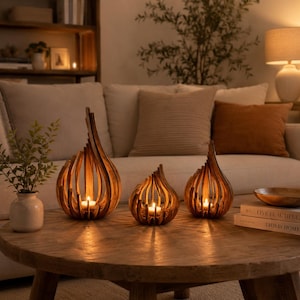 Peut inclure: Trois bougeoirs en bois au design incurvé en forme de flamme, chacun contenant une petite bougie chauffe-plat allumée. Les bougeoirs sont disposés sur une table ronde en bois, le plus grand à gauche et deux plus petits à droite.
