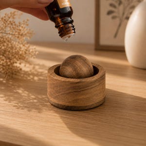 Peut inclure: Une petite sphère en bois brun foncé, ronde, reposant à l'intérieur d'une base cylindrique en bois assortie. Un flacon compte-gouttes en verre ambré foncé est placé au-dessus de la sphère, avec une goutte de liquide clair suspendue.