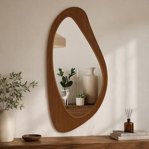 Peut inclure: Un miroir mural asymétrique doté d'un cadre en bois foncé lisse. Le miroir présente une forme organique irrégulière qui reflète l'intérieur d'une pièce contenant un grand vase en céramique, une petite plante en pot, une figurine de cerf et des bougies.