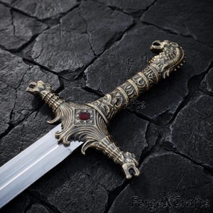 May include: A decorative sword featuring an ornate bronze-toned metal hilt with intricate relief carvings, a pommel shaped like a dragon head, and a crossguard centred with a rectangular red stone. The blade is polished silver-toned steel.