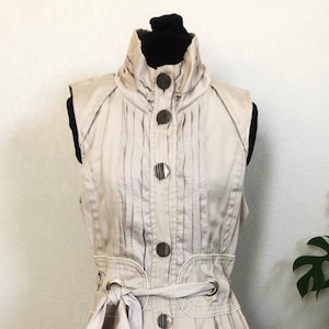 May include: A sleeveless beige dress with a high collar, button closure, and a matching belt. The dress has a pleated bodice and a slightly flared skirt.