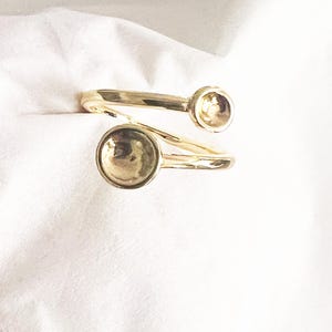 May include: A gold-colored ring with a unique design. The ring features two circular, raised elements on a spiral band. The ring is set against a white background, highlighting its metallic sheen and intricate form.