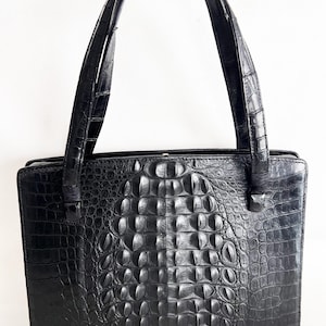 May include: A black handbag with a crocodile skin pattern. The bag has two handles and a rectangular shape. The surface of the bag is textured, and the color is a deep black. The bag appears to be made of leather or a similar material.