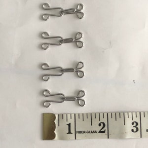 May include: Four silver metal hook and eye closures, each measuring approximately 1 inch in length, are lined up on a white surface. A ruler with the numbers 1, 2, and 3 is visible below the closures.