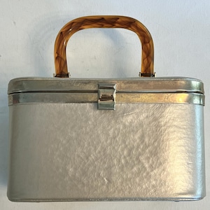 May include: A vintage silver-colored handbag with a textured surface and a curved, brown tortoiseshell handle. The rectangular bag has a silver metal frame and clasp, suggesting a classic, retro design.