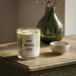 Könnte beinhalten: Eine zylindrische Glaskerze mit einer geschichteten Optik, die an einen Eismatcha-Latte erinnert, mit einem weißen Etikett mit der Aufschrift Pure Candle Iced Matcha Latte, einem zentralen Docht und hellgrünem Wachs mit klaren Wachswürfeln.
