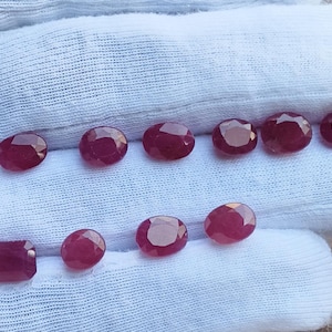 May include: A collection of ten loose, faceted rubies in deep red hues. Nine of the gemstones are oval-shaped, while one is rectangular. The stones are displayed on a white fabric surface, showcasing their polished surfaces and light-reflecting facets.