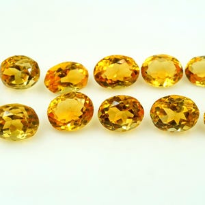 May include: A collection of eleven faceted citrine gemstones, each cut into a uniform oval shape. These stones exhibit a vibrant, translucent golden-yellow hue with internal light reflections visible across their polished, multi-faceted surfaces.