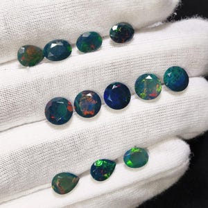 May include: A collection of twelve faceted loose gemstones, primarily dark blue and black with iridescent flashes of green, red, and orange. The stones are arranged in three rows on a white fabric surface, featuring a mix of round, oval, and pear shapes.
