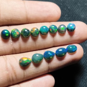 May include: A collection of thirteen small, smooth, oval-shaped black opals arranged in two rows. Each gemstone features a dark base color with iridescent flashes of green, blue, orange, and red light reflecting from within the polished surfaces.