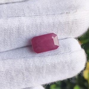 May include: A rectangular, faceted gemstone with a deep magenta-pink hue. The stone features a classic emerald-cut shape with beveled edges, displaying a smooth, polished surface that reflects light across its saturated, uniform color profile.