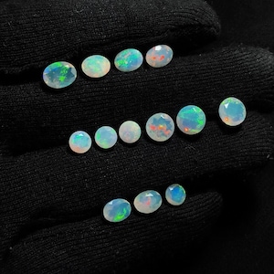 May include: A collection of thirteen faceted, translucent white opals arranged in three rows. Each gemstone displays a play-of-color with flashes of green, blue, and orange. The stones are oval and round in shape, showcasing a polished surface.