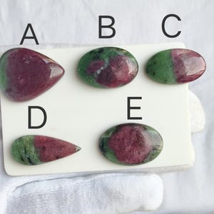 Puede incluir: Cinco cabujones de rubí en zoisita etiquetados de la A a la E sobre una tarjeta blanca. Cada piedra presenta una combinación de inclusiones de rubí rojo magenta intenso en una matriz de zoisita verde vibrante, con superficies pulidas.