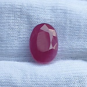 May include: A vibrant, oval-shaped ruby gemstone featuring a faceted surface that reflects light. The deep red stone displays a polished finish with distinct geometric cuts across its top, showcasing a rich, saturated colour and a clear, translucent quality.