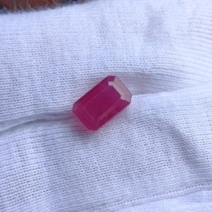 May include: A vibrant, deep pinkish-red gemstone cut into a rectangular emerald shape with faceted edges. The stone displays a translucent quality and sits against a white, textured fabric background, highlighting its geometric form and color.