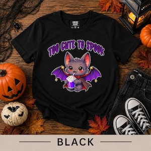 May include: A black short-sleeve t-shirt featuring a graphic of a grey cartoon bat with purple wings, wearing a red cape and holding a purple drink. Above the bat, the text reads TOO CUTE TO SPOOK in purple letters with a white outline.