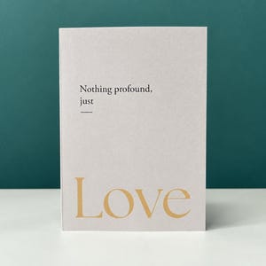 Może przedstawiać: Prostokątna kartka okolicznościowa z jasnoszarym tłem. Tekst "Nothing profound, just" jest wydrukowany na czarno, a pod nim znajduje się pozioma linia. Poniżej słowo "Love" jest wydrukowane dużą czcionką szeryfową w kolorze złotym.