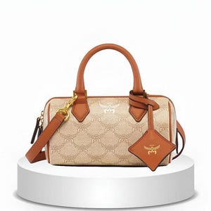 Könnte beinhalten: Eine beige-braune, zylinderförmige Handtasche mit einem sich wiederholenden Monogramm-Muster. Die Tasche verfügt über hellbraune Ledergriffe, einen abnehmbaren Schulterriemen mit goldfarbenen Beschlägen und einen rautenförmigen Lederanhänger.