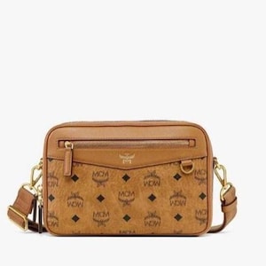 Könnte beinhalten: Eine rechteckige Umhängetasche in Cognac mit schwarzem MCM-Monogramm-Aufdruck. Die Tasche verfügt über eine vordere Reißverschlusstasche, einen D-Ring aus goldfarbenem Metall, einen Reißverschluss oben und einen verstellbaren Schulterriemen mit goldfarbenen Beschlägen.