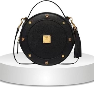 Könnte beinhalten: Eine schwarze, runde Handtasche aus Leder mit einer goldfarbenen Metallplakette mit dem MCM-Logo in der Mitte. Die Tasche ist mit goldfarbenen Nieten und kleinen Logo-Emblemen am Rand, einem Tragegriff, einem Schulterriemen und einer Quaste verziert.