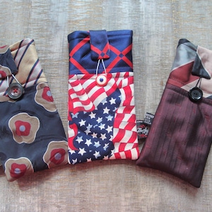May include: Three handmade phone cases crafted from repurposed neckties. The cases feature various patterns, including stripes, floral designs, and an American flag motif. Each case has a button closure and a small tag.
